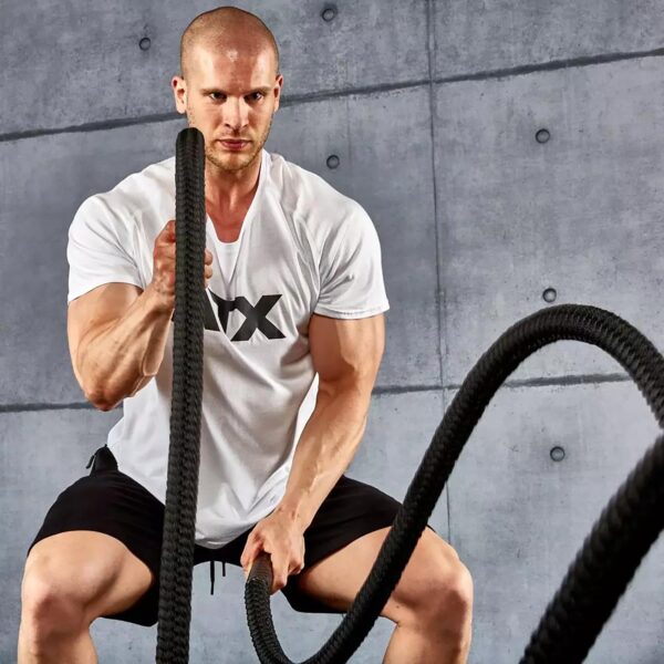 A man in an ATX® white t-shirt and black shorts works out with battle ropes in a gym, crouched in an athletic stance against a concrete wall, showcasing strength and focus.