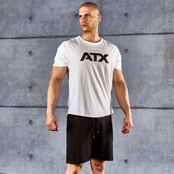 A man with a shaved head stands in front of a concrete wall, wearing an ATX® White T-shirt and black shorts. He looks to the side with a neutral expression.
