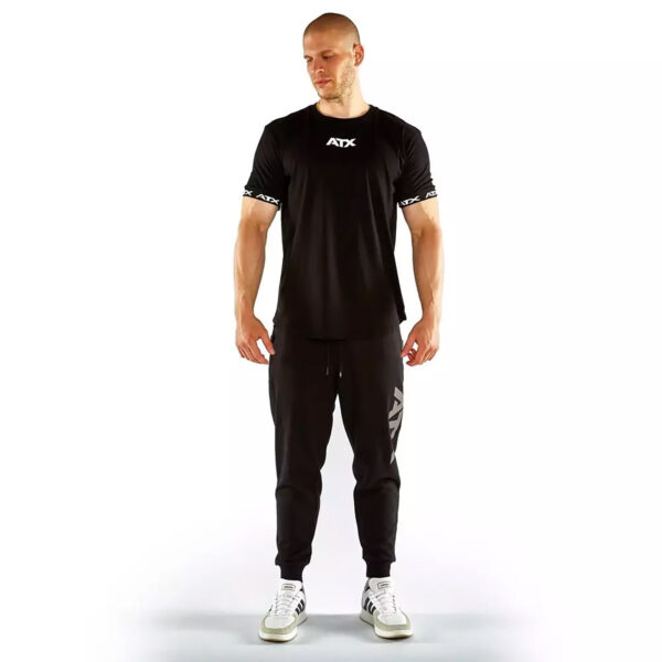 A man in a black ATX® Training Shirt BLACK, black joggers, and white sneakers stands against a plain white background. Hes bald with downcast eyes.