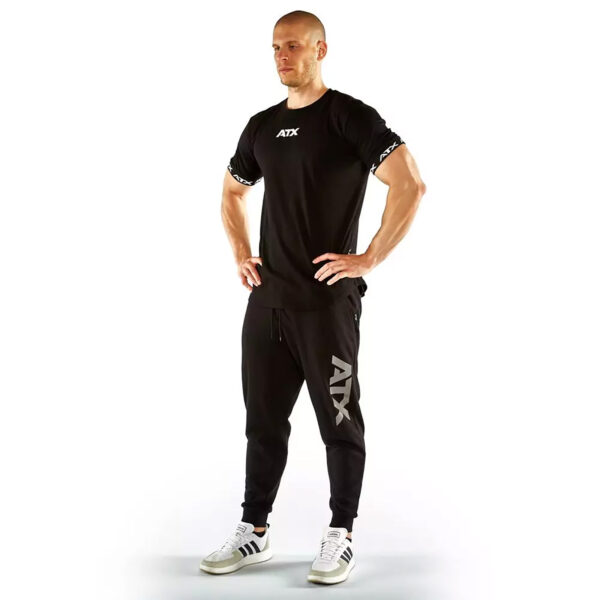 A person stands confidently with hands on hips, wearing a black ATX® Training Shirt and matching joggers, complemented by white sneakers against a plain white background.