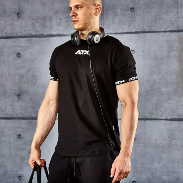 A person in an ATX® Training Shirt BLACK and black pants stands holding a bag with headphones around their neck, set against a gray, textured wall. Their head is slightly turned to the side.