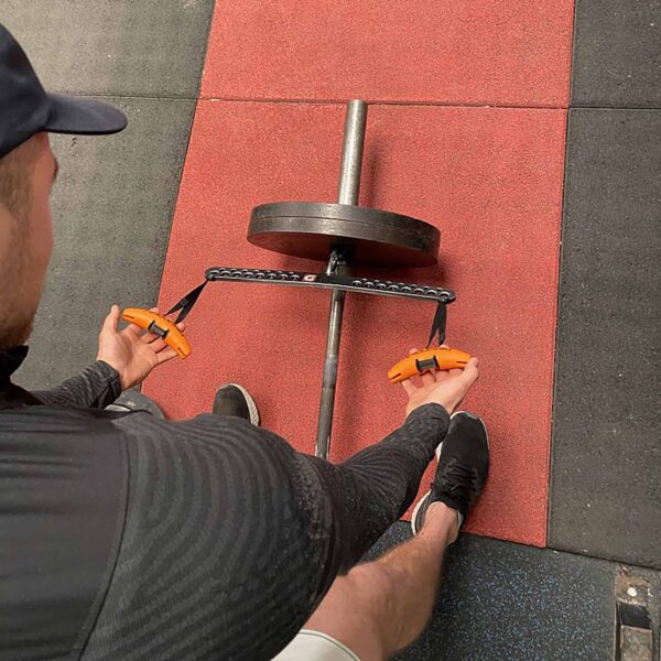 A person in athletic wear grips two orange T-BAR Row attachment Collars connected by a black chain, standing over a weightlifting bar with a plate, poised to lift on a gym floor with black and red tiles.