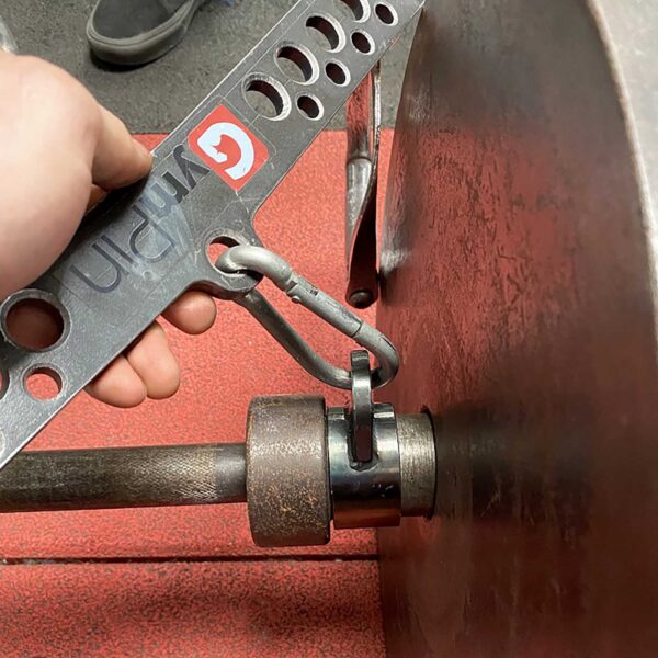 Close-up of a gym weightlifting setup. A hand holds the T-BAR Row attachment Collar connected to a barbell with weight plates. The barbell is secured by a clip on a red gym floor, and another pair of shoes is visible in the background.