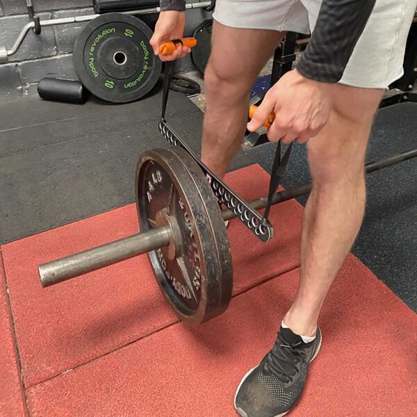 In a gym setting, someone in shorts and sneakers uses a hacksaw to cut a weightlifting bar with the T-BAR Row attachment Collar and weights in place, while another barbell with attached weights lies on the ground nearby.