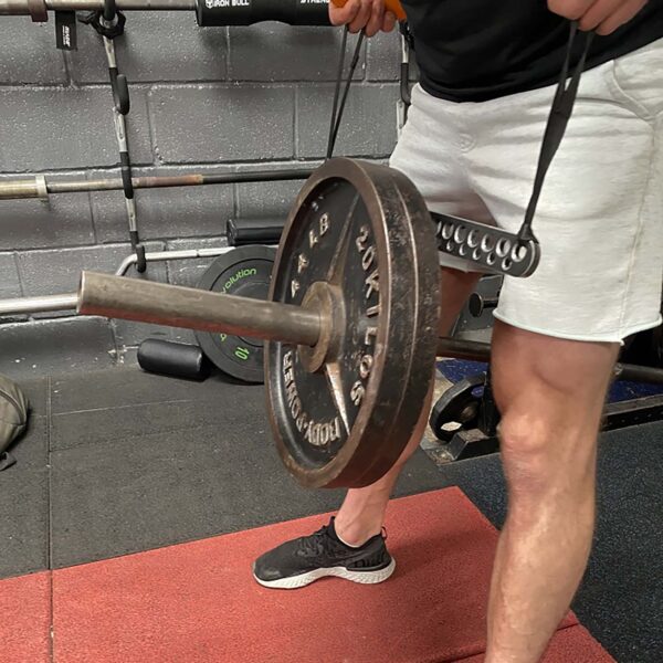 A person using a T-BAR Row attachment Collar with a weight plate, standing on a gym mat amid weightlifting bars and plates. They are wearing black shoes and white shorts.