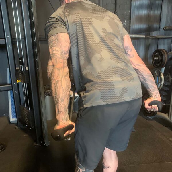 A person with tattoos, in a patterned shirt and shorts, is lifting TriGrips Long Handles in a gym filled with various weights and equipment.