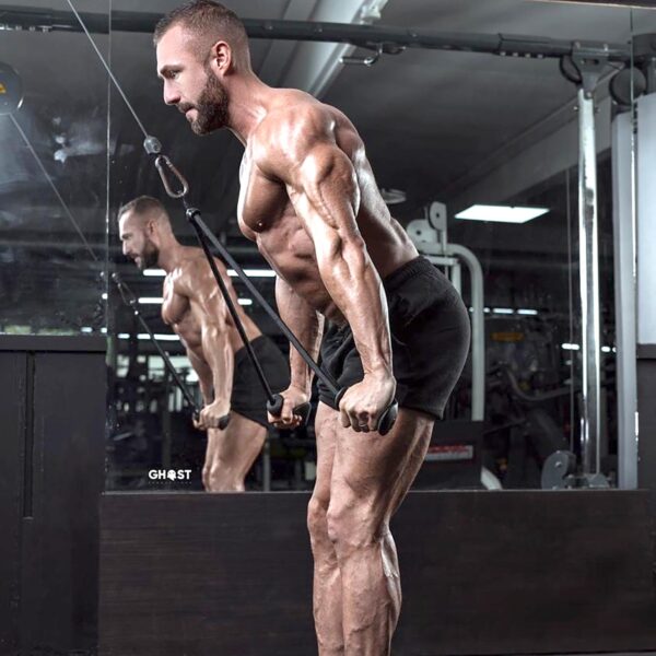 A muscular man is using TriGrips Long Handles for a triceps pushdown on a cable machine. He is shirtless, in black shorts, at a gym with mirrors reflecting his image.