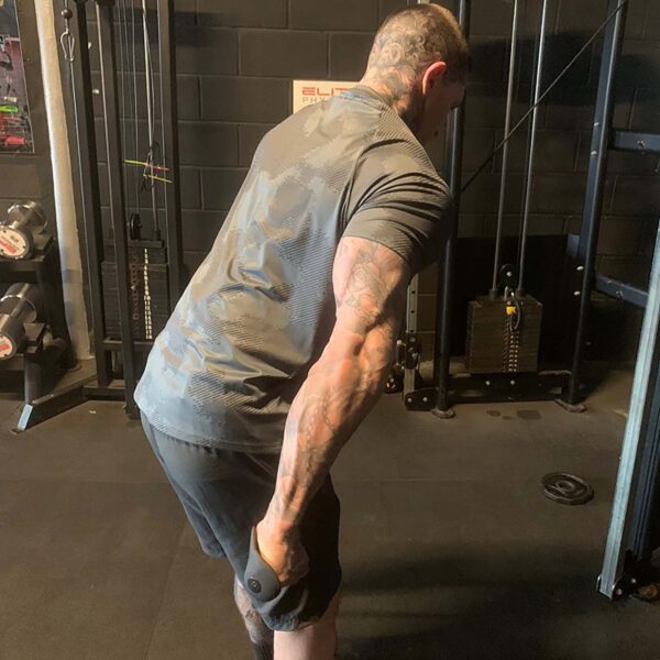 A tattooed person is performing a tricep exercise in a gym with TriGrips Long Handles, wearing a gray shirt and black shorts, amidst visible gym equipment and weights.