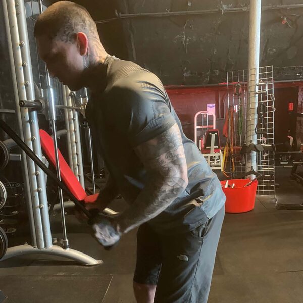 A tattooed person is working out in a dimly lit gym, using TriGrips Long Handles for tricep exercises. Theyre wearing a gray shirt and black shorts, surrounded by gym equipment like a bench and weight rack.