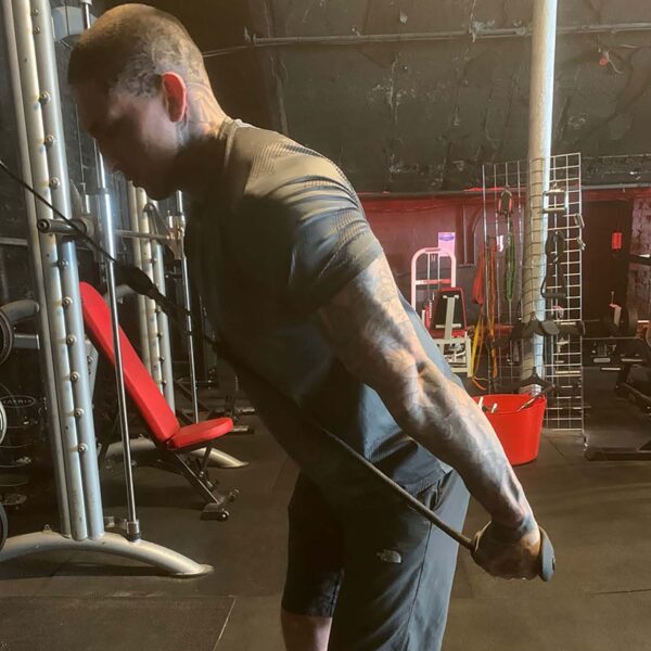 A tattooed individual exercises in a gym, using TriGrips Long Handles on a cable machine for tricep pushdowns. The gym features diverse equipment like a red bench and weights, all set against dark floors and ceilings.