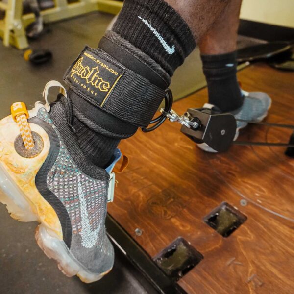 A person in black Nike socks and sneakers, wearing Spud Ankle Cuff Pair connected to a pulley system for leg exercises in a gym with a wooden floor that has a visible hole.