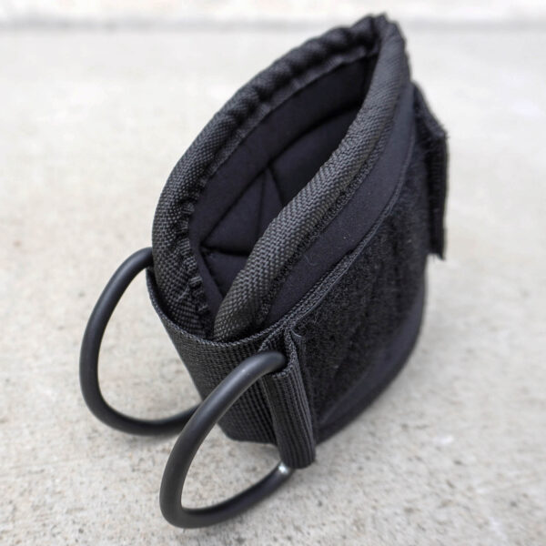 The Spud Ankle Cuff Pair features a close-up of a black fabric strap with two D-rings on a concrete surface, showcasing visible stitching and a quilted pattern on one side, complemented by Velcro material on the other.
