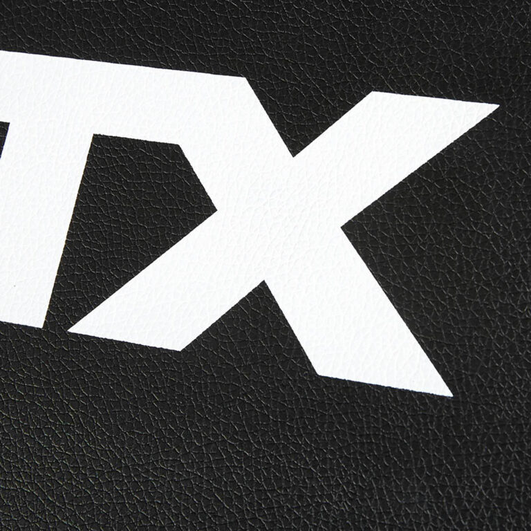 Close-up of a white, bold, partial letter X on a textured black surface, reminiscent of leather, as if carved into the robust material of a fid bench.