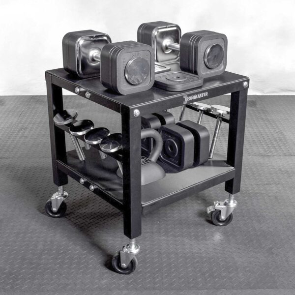 On a textured gray floor, the black Ironmaster QLDB Stand PRO holds various gym equipment: Ironmaster adjustable dumbbells on top, cylindrical dumbbells in the middle, and kettlebells on the bottom shelf.