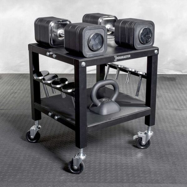The Ironmaster QLDB Stand PRO, a black wheeled rack, neatly holds adjustable dumbbells and a kettlebell on a textured gray floor. It has extra weights and includes a compartment for weight plate storage.