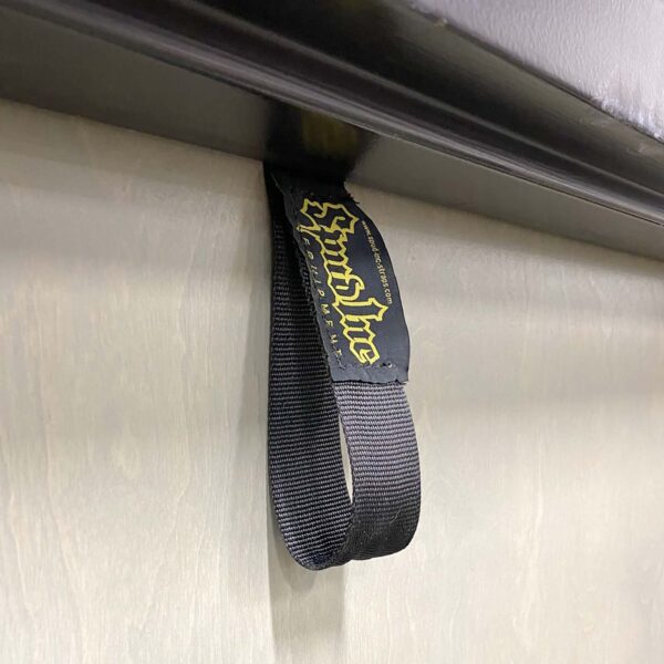 Close-up of a black Spud Inc Door Jammer strap featuring Sound Line in yellow lettering, attached to a horizontal metallic surface, set against a textured wall.