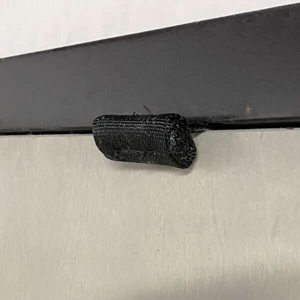 Close-up of a black Velcro strap from the Spud Inc Door Jammer attached to a two-toned metal surface, secured with a metal bracket. The fabric at the edges appears slightly frayed.