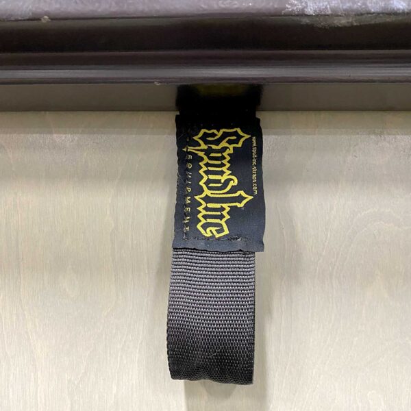 A black fabric strap with a gold and white Grindline and Dreamland logo, attached to a wooden surface. Product: Spud Inc Door Jammer.