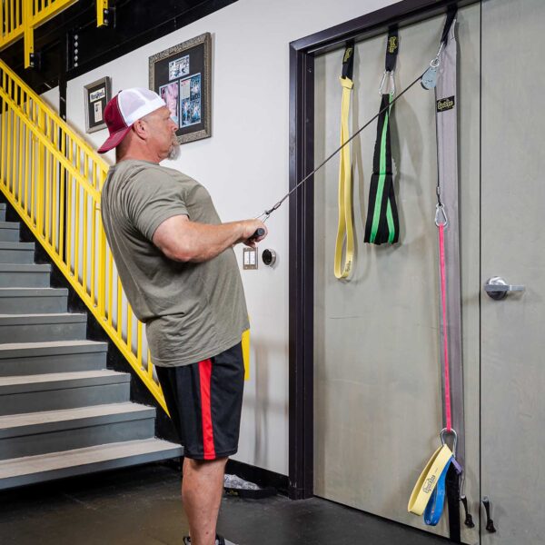 A man in workout attire exercises with the Spud Inc Door Jammer Pulley System attached to a door. He pulls the bands towards him at an angle, with a yellow staircase and a framed picture visible in the background.