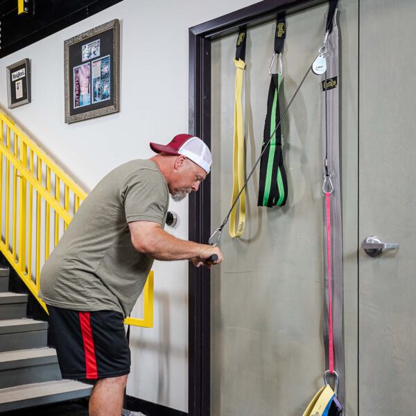 A man in a backward baseball cap and athletic wear exercises with the Spud Inc Door Jammer Pulley System attached to a door. Hes near a staircase with yellow railings, and framed pictures adorn the wall.