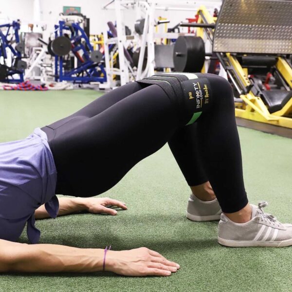 A person uses the Spud Inc Infinity Loop for a bridge exercise on a gym mat. Theyre in black leggings and gray sneakers, with gym equipment visible in the background.