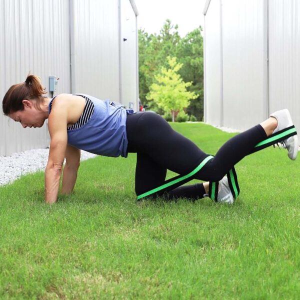 A woman exercises outdoors on grass between buildings. She kneels, wearing a blue tank top and black leggings, using the Spud Inc Infinity Loop around her thighs. She extends one leg backward, focusing on stability and strength.