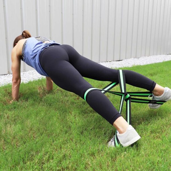 A person in athletic gear performs a plank on grass using Spud Inc Infinity Loop green resistance bands around their legs, near a metal fence.