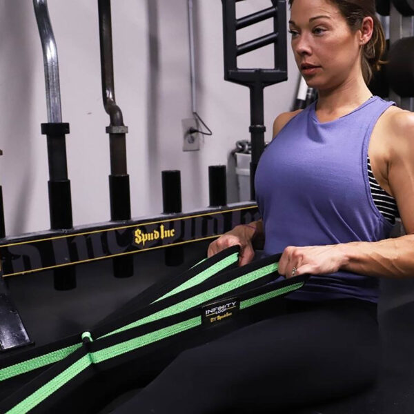 A woman in a purple tank top sits on the gym floor, using a green Spud Inc Infinity Loop for resistance exercises. She is focused and performing a seated pull exercise with gym equipment in the background.