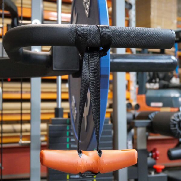 Close-up of a gym setup featuring a black bar attached to a metal frame, with a T-shaped orange D Angle Grip V2 connected by black straps. Weight plates and other gym apparatus are visible in the background.