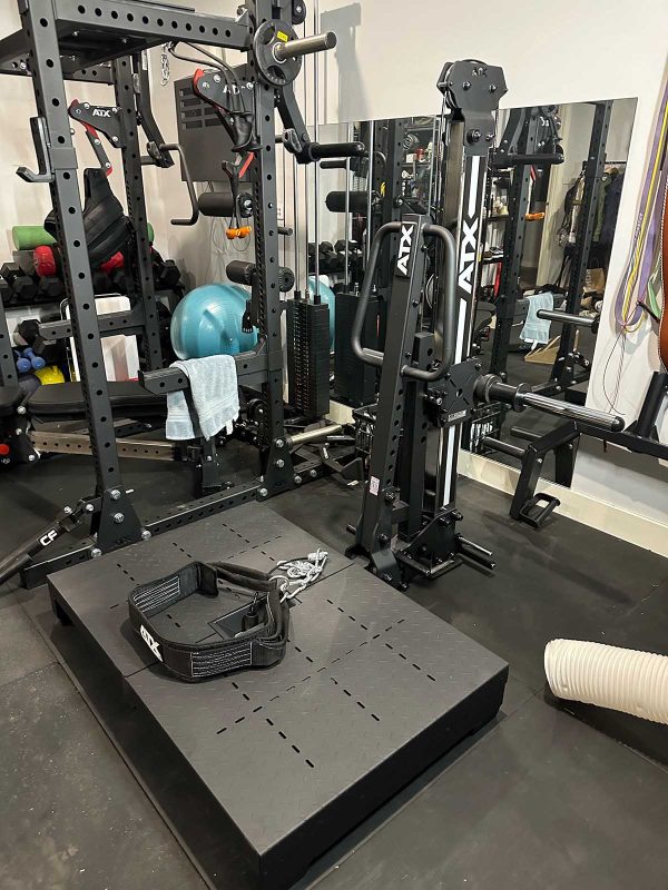 A gym setup with various exercise equipment, including a power rack, resistance bands, exercise balls, and a platform with a handle attachment. The area is organized and surrounded by mirrors.