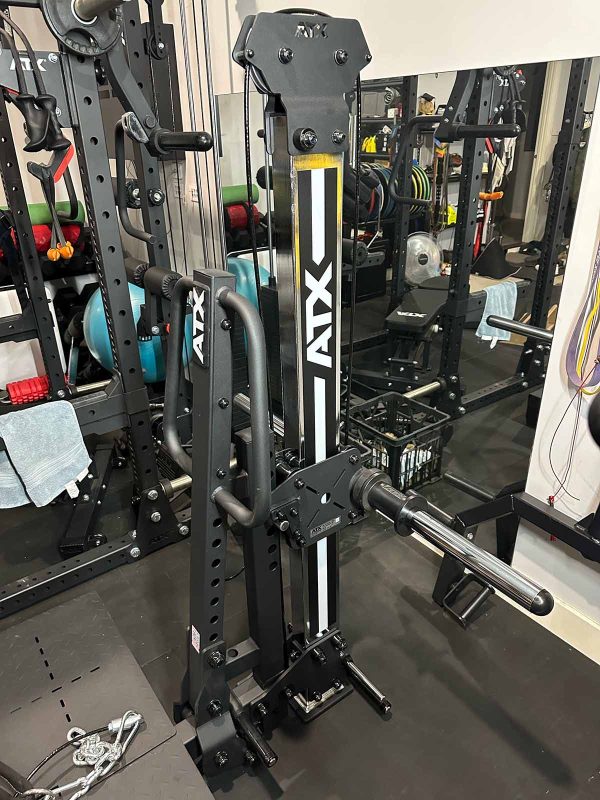 A black ATX fitness machine in a gym with various workout equipment in the background, including free weights and exercise balls. The machine features metal bars and adjustable components for strength training.