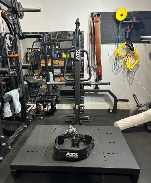 A home gym setup with various exercise equipment including a squat rack, barbells, and belts. A workout belt is placed on a platform in the foreground, and resistance bands hang on the wall. A mirror reflects the equipment.