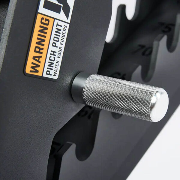 Close-up of a metal handle with textured grip on a black weightlifting machine near the ATX® Commercial FID Bench ATX-MBX-660. It has a warning label: Pinch Point, Watch Your Fingers. Part of the numbered weight stack is visible.