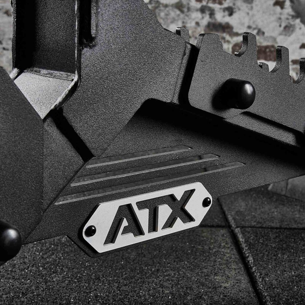 Close-up of a black metal gym equipment piece, featuring a silver plate displaying the letters ATX. An adjustable weight bench forms a textured surface in the background.