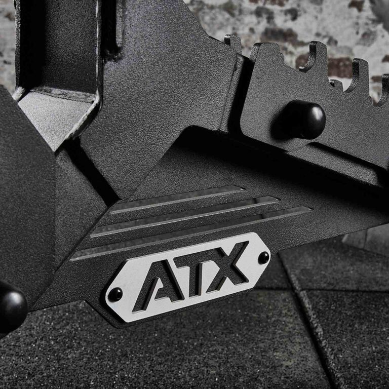 Close-up of a black metal gym equipment piece, featuring a silver plate displaying the letters ATX. An adjustable weight bench forms a textured surface in the background.