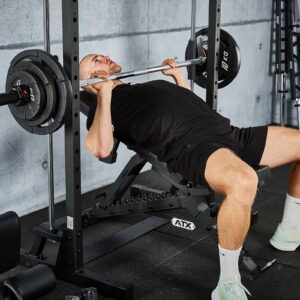 In a gym with concrete walls, a person in black is using the ATX® Smith Machine 500 (198 cm) for bench pressing, surrounded by fitness equipment.