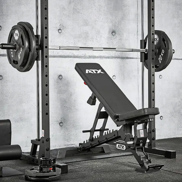 An ATX® Smith Machine 500 is set with a black adjustable weight bench under the barbell, loaded with 20 kg plates against a concrete wall. A small plate lies on the floor nearby.