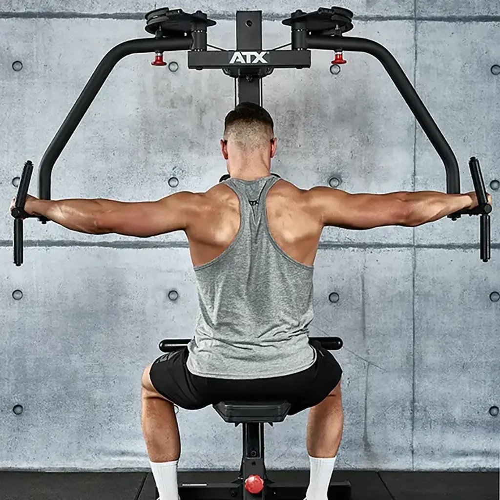 A person wearing a gray tank top and black shorts is using a pectoral fly machine in a gym. Seated and facing a concrete wall, they extend their arms outward. Fitness equipment surrounds them, highlighting a focused workout environment.