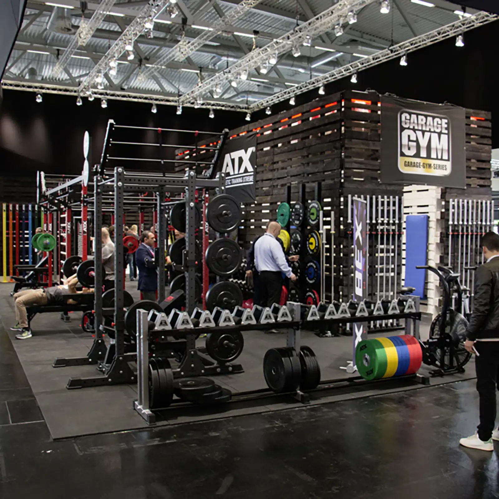 The gym area is well-equipped with various fitness machines and weights. Several people exercise using equipment like the ATX® Warrior Bench 2.0, barbells, and a stationary bike. Industrial lighting enhances the atmosphere.