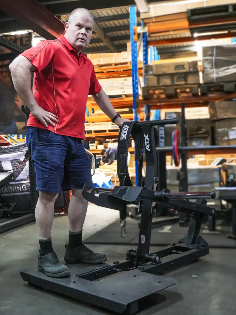 A man in a red shirt and blue shorts stands beside the ATX® Belt Squat Compact in a warehouse lined with shelves. He has one hand on his hip and is wearing work boots.