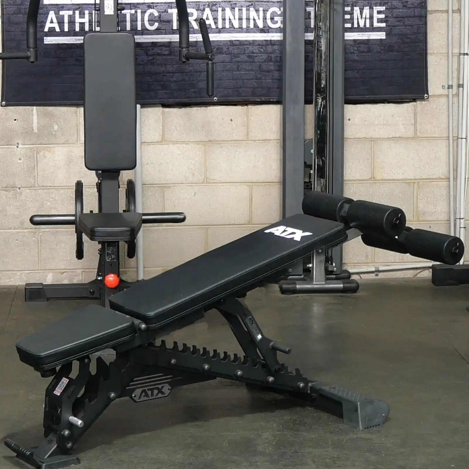 An ATX® Warrior Bench 2.0 with a padded seat and leg supports is prominently in the foreground, while a workout machine and a Athletic Training sign are partially visible in the background.