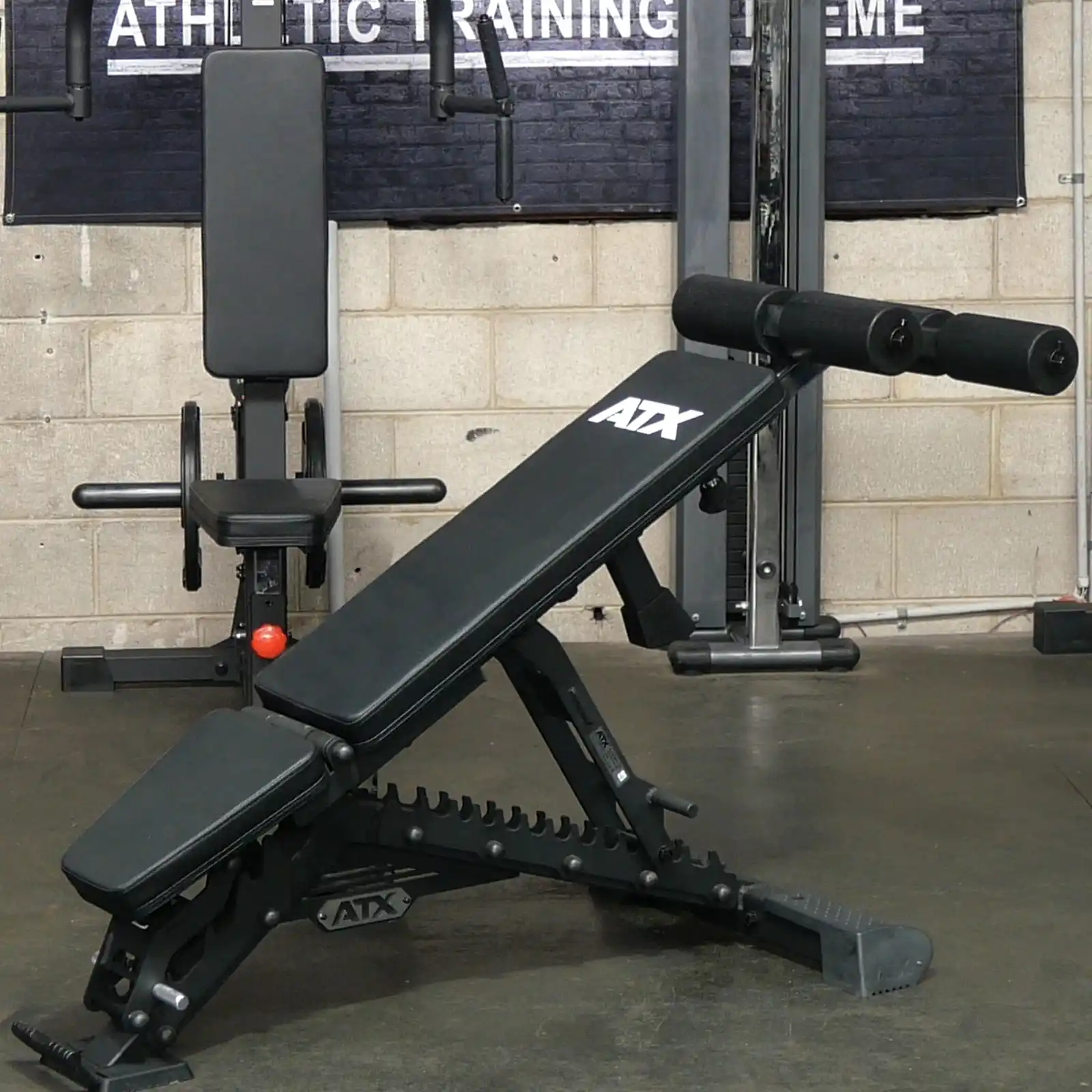 An ATX® Warrior Bench 2.0 with a black design and logo on the backrest is set in a workout area, backed by a beige brick wall and assorted gym equipment, crafting an authentic fitness scene.