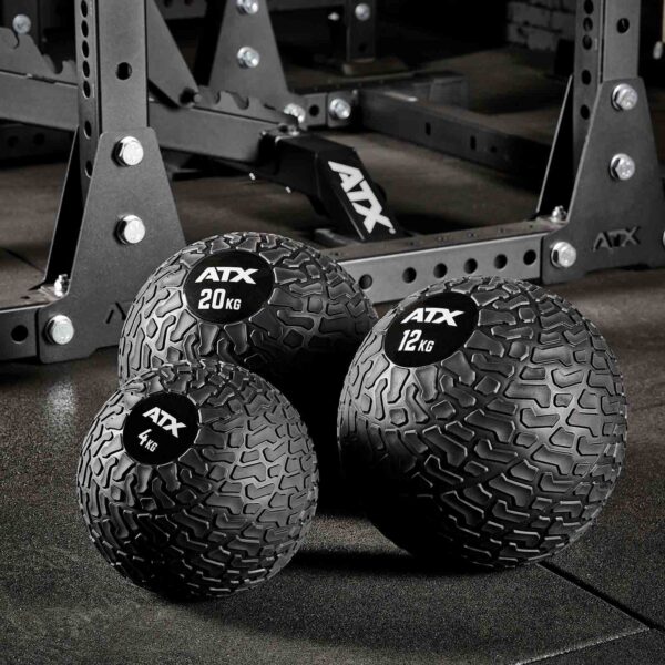 Three ATX® Slam Balls in black texture (4 kg, 12 kg, 20 kg) rest on the gym floor with a metal exercise rack seen behind.