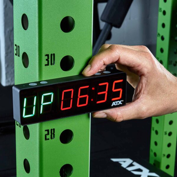 A person adjusts a digital timer attached to a green gym rack. The display shows UP in green and 06:35 in red. The rack features multiple numbered holes for adjusting height.