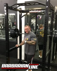A man in a gym stands next to a squat rack with heavy weights, giving a thumbs up. Hes wearing a gray T-shirt and black shorts. The gym has various equipment, and a logo reading “Barbarian Line” is visible on the rack and in the foreground.