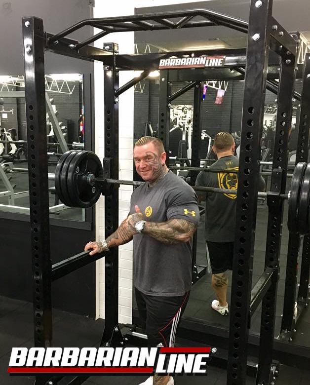 A man in a gym stands next to a squat rack with heavy weights, giving a thumbs up. Hes wearing a gray T-shirt and black shorts. The gym has various equipment, and a logo reading “Barbarian Line” is visible on the rack and in the foreground.