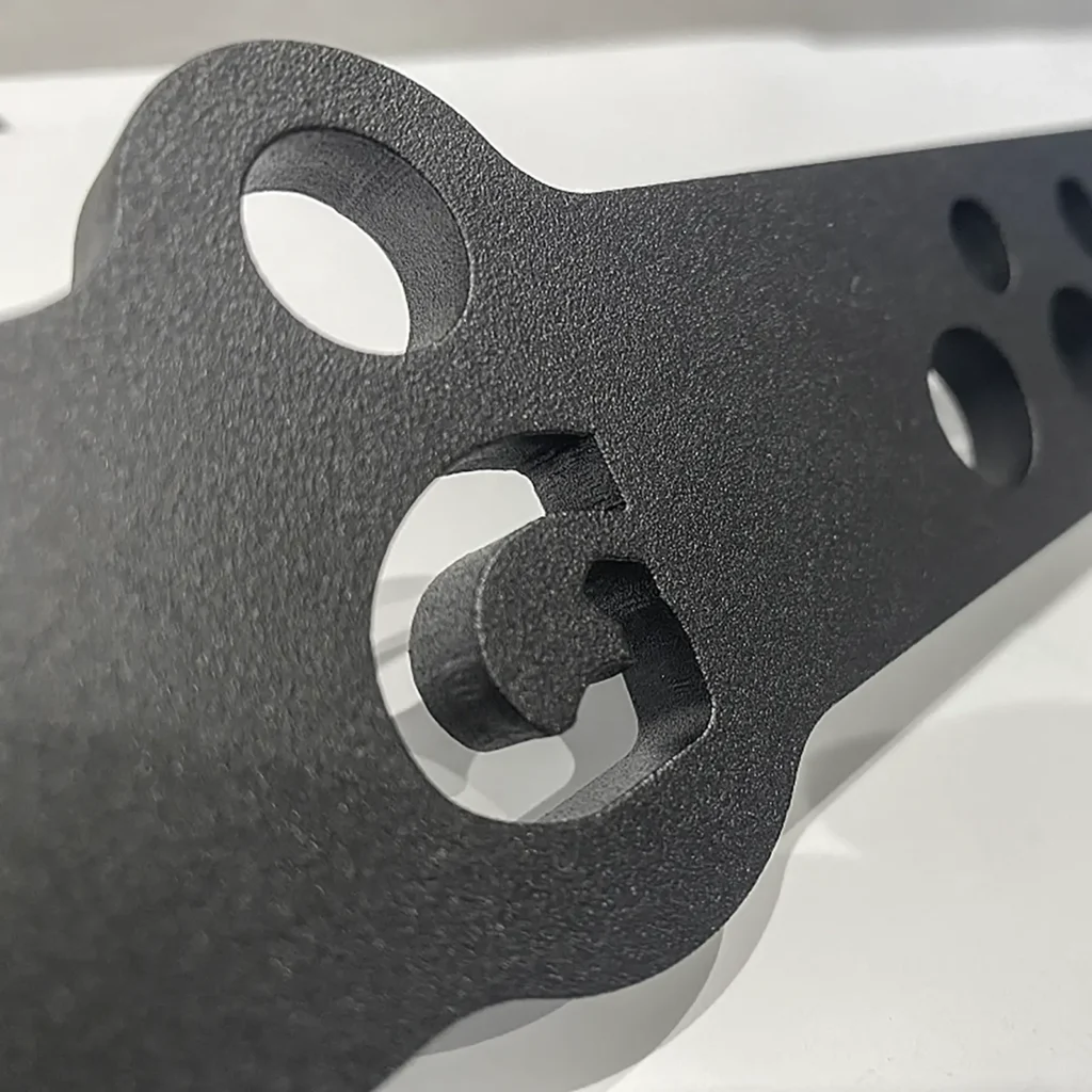 Close-up of a GymPin Lite 20inch D Bar, featuring black metal with multiple holes and a textured surface, resting on a reflective white base. The image highlights intricate cutouts and contoured edges, suggesting its industrial or mechanical application.