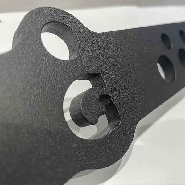 Close-up of a GymPin Lite 20inch D Bar, featuring black metal with multiple holes and a textured surface, resting on a reflective white base. The image highlights intricate cutouts and contoured edges, suggesting its industrial or mechanical application.