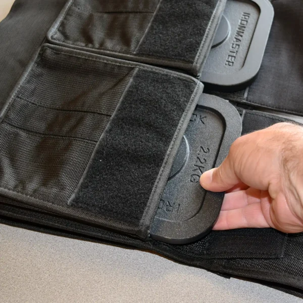 A hand inserts a weight plate, embossed with IRONMASTER and 2.2 KG, into a black fabric pocket featuring Velcro closures. This pocket is part of the Ironmaster Ultimate Training Vest, which rests on a light gray surface, enhancing the functionality of this efficient fitness vest.