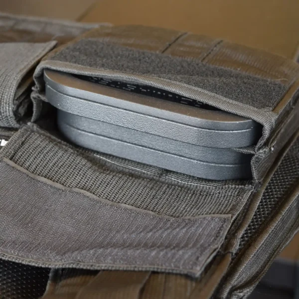 A close-up of the Ironmaster Ultimate Training Vest pocket shows a stack of three rectangular, black trauma pads. The partially open pocket reveals the pads, secured with Velcro straps. The vest features a dark gray fabric with a textured surface.